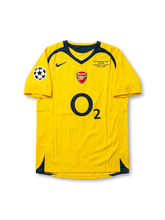 Front view of Arsenal 2006 Champions League Final away shirt with yellow body, O2 sponsor, Arsenal crest, Paris final match detail, and UEFA sleeve patch.