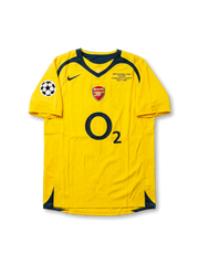 Front view of Arsenal 2006 Champions League Final away shirt with yellow body, O2 sponsor, Arsenal crest, Paris final match detail, and UEFA sleeve patch.