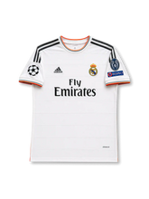 Front view of Real Madrid 2013-14 home shirt with white body, Fly Emirates sponsor, Real Madrid crest, grey shoulder stripes, orange trim, and UEFA Champions League sleeve patches.