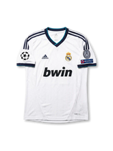 Front view of Real Madrid 2012-13 home jersey with white base, navy shoulder stripes, teal trim, club crest, sponsor, and Champions League sleeve patches.