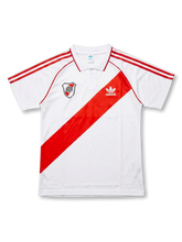 Front view of River Plate retro sash polo shirt with white base, red diagonal sash, club crest, red shoulder stripes, and structured polo collar.