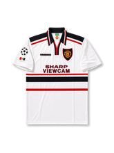 Front view of Manchester United 1997-99 away shirt with white base, black collar, red and black chest stripes, club crest, Sharp Viewcam sponsor, and Champions League sleeve patch.