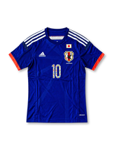日本代表 2018年 ホームユニフォーム キャプテン翼10番 スペシャルエディション。濃いジャパンブルーのボディに刺し子風の縦ライン柄、ホワイトの肩のスリーストライプ、レッドの縁取りを施したVネックを組み合わせ、胸元に日本国旗、JFAエンブレム、adidasロゴ、キャプテン翼の漫画イラスト入り10番を配したサッカージャージ。両袖にはキャプテン翼とLiving Football FIFAのデザインパッチをあしらった特別仕様。