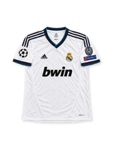 レアル・マドリード 2012/13シーズン ホームユニフォーム アンヘル・ディ・マリア22番。adidas製ホワイトのボディに細かな縦ストライプの織り柄、ネイビーの肩のスリーストライプと袖口、ターコイズブルーの縁取りを施したVネックを組み合わせたレトロサッカージャージ。胸元にはadidasロゴ、レアル・マドリードのクラブエンブレム、ブラックのbwinスポンサーロゴを配置。UEFAチャンピオンズリーグ仕様の袖章を備え、背面にDI MARIAのネームと22番を配したデザイン。