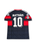 バイエルン・ミュンヘン 1997〜1999年 アウェイユニフォーム ローター・マテウス10番。adidas製のダークネイビーのボディにレッドの太い胸ラインと袖の切り替え、ホワイトの折り襟と肩のスリーストライプを組み合わせたレトロサッカージャージ。胸元にはadidasロゴ、バイエルンのクラブエンブレム、ホワイトのOPELスポンサーロゴを配置。UEFAチャンピオンズリーグの袖章を備え、背面にはMATTHÄUSのネームとホワイトの10番を配したデザイン。