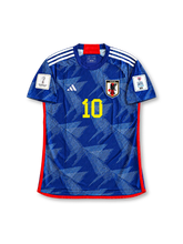 日本代表 2022年カタールワールドカップ ホームユニフォーム 南野拓実10番。adidas製ジャパンブルーのボディに折り紙と折り鶴をモチーフにした幾何学模様を施し、ホワイトの肩のスリーストライプとレッドのサイドアクセントを組み合わせたサッカージャージ。胸元にadidasロゴ、JFAエンブレム、イエローの10番を配置。袖にはカタールワールドカップとFootball Unites the Worldのデザインパッチを備え、背面には日本国旗、MINAMINOのネームとイエローの10番を配したモデル。