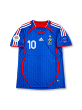フランス代表 2006年ドイツワールドカップ ホームユニフォーム ジネディーヌ・ジダン10番。adidas製のロイヤルブルーのボディにレッドのVネック、ホワイトとレッドの肩のスリーストライプ、脇から裾へ続くレッドの曲線パネルを組み合わせたレトロサッカージャージ。胸元にはホワイトのadidasロゴ、ゴールドの星付きフランスサッカー連盟エンブレム、ホワイトの10番を配置。袖には2006年ワールドカップの大会デザインパッチを備え、背面にはZIDANEのネームとホワイトの10番を配したデザイン。