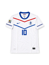 オランダ代表 2010年南アフリカワールドカップ アウェイユニフォーム ウェスレイ・スナイデル10番。Nike製のホワイトのボディにブルーのクルーネック、胸元のレッドとブルーのV字型グラフィック、袖口のブルーのラインを組み合わせたレトロサッカージャージ。胸元にはブルーのNikeロゴ、KNVBエンブレム、ブルーの10番を配置。袖には2010年ワールドカップの大会パッチを備え、背面にはSNEIJDERのネームとブルーのドット柄10番、襟下にはNEDERLANDの文字をあしらったデザイン。