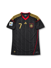 Germany 2010 away jersey featuring Bastian Schweinsteiger #7, adidas black shirt with gold three-stripe shoulder detailing, red V-neck collar, gold adidas logo, three-star Germany national team crest, and SCHWEINSTEIGER name and number 7 on the back, inspired by the 2010 FIFA World Cup in South Africa.