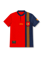Spain 1996 home jersey with a red body, asymmetric navy panel, narrow red-and-yellow vertical stripes, navy button collar, gold adidas logo, Spanish federation crest and sleeve flag.