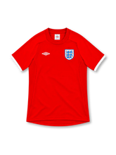 England 2010 red Umbro away jersey with a rounded crew-neck collar, white sleeve cuffs, white double-diamond Umbro logo, Three Lions crest, raglan shoulder seams and curved hem.