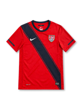 This red shirt features a wide navy sash running diagonally from the shoulder to the opposite hem, edged on both sides by thin navy pinstripes, along with a navy crew-neck collar and navy bands on both sleeves. The white Nike swoosh sits on the sash on the right chest opposite the embroidered U.S. Soccer shield crest with three stars, and a Dri-Fit logo and Nike Authentic tag appear near the lower hem, with navy taping inside the neck.