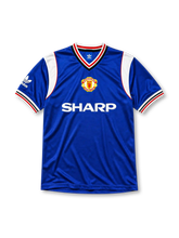 Manchester United 1985-86 third jersey, front view, royal blue body, white striped V-neck collar, white sleeve panels with black and red trim, adidas trefoil logo on sleeve, club crest, SHARP sponsor logo, striped ribbed cuffs