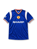 Manchester United 1985-86 third jersey, front view, royal blue body, white striped V-neck collar, white sleeve panels with black and red trim, adidas trefoil logo on sleeve, club crest, SHARP sponsor logo, striped ribbed cuffs