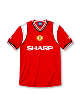 Manchester United 1984-86 home jersey, front view, red body, white sleeve panels with black and red trim, red white and black striped V-neck collar, Umbro diamond logo on sleeves, club crest, SHARP sponsor logo, striped ribbed cuffs