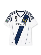 LA Galaxy 2012 home jersey, front view, white body with navy diagonal diamond-pattern sash, adidas three-stripe shoulder detailing, MLS Cup Champion patch, MLS league patch, club crest, Herbalife sponsor logo