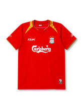 Liverpool 2005-06 home jersey, front view, red body with gold shoulder panels and collar trim, V-neck collar, white Reebok logo, Liverpool crest with five stars, Carlsberg sponsor logo, small winner's patch near bottom hem