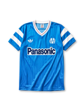 Marseille 1990 away jersey, front view, sky blue body with tonal pinstripe pattern, white V-neck collar with red and blue piping, white adidas trefoil logo, circular Olympique de Marseille crest, navy Panasonic sponsor logo