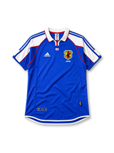 Japan 2000 home jersey, front view, blue body with white shoulder panels and red trim, white polo-style collar with red zip placket, small Japan flag emblem, JFA crest with three-legged crow, Japan text beneath crest