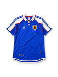Japan 2000 home jersey, front view, blue body with white shoulder panels and red trim, white polo-style collar with red zip placket, small Japan flag emblem, JFA crest with three-legged crow, Japan text beneath crest