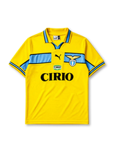 Lazio 1998-99 away jersey, front view, bright yellow body with sky blue chest band, yellow polo-style collar with black and blue trim, black Puma logo, S.S. Lazio eagle crest, Cirio sponsor logo and badge, sky blue cuffs