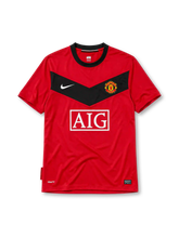 Manchester United 2010 home jersey, front view, red body with black chevron panel, black crew-neck collar, white Nike swoosh, Manchester United crest, boxed white AIG sponsor logo