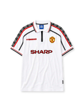 Manchester United 1998-99 away jersey, front view, white body with red and black diamond pattern shoulder trim, zip-up polo collar, red Umbro logo, Manchester United crest, red Sharp sponsor logo