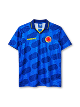 Colombia 1994 away jersey, front view, royal blue body with tonal diamond pattern and darker football-panel print, navy polo collar with yellow tipping and V insert, yellow UMBRO wordmark on right chest, Colombian Football Federation crest on left chest