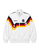 Front view of a white Germany 1990 World Cup retro track jacket featuring black, red, and gold chevron graphics across the chest and sleeves, a black adidas Trefoil logo, circular DFB crest, stand-up collar, full zipper, and elasticated cuffs.