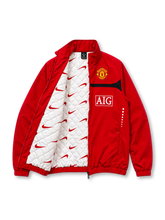Front view of the Manchester United 2009/10 Nike red track jacket featuring a curved black chest stripe, white Nike Swoosh, Manchester United club crest, AIG sponsor logo, high collar, full-length front zipper, and two zippered pockets.