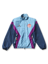 Paris Saint-Germain × Jordan Brand track jacket in ice blue with diagonal purple chevron panels, Jumpman logo at left chest, coral PSG crest badge at right chest, navy sleeves