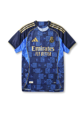 Real Madrid × adidas × Louis Vuitton away jersey in navy with LV geometric monogram patchwork print, sky blue side panels, gold Three Stripes on shoulders, gold adidas logo at left chest, gold Real Madrid crest at right chest, Emirates gold sponsor