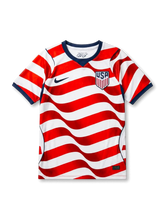 USA national team Nike 2026/27 home jersey in white with full-body wavy red and white stripe pattern, navy V-neck collar, navy curved side panels, navy Swoosh at left chest, USA shield crest at right chest, navy sleeve cuff bands