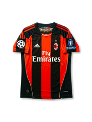 Front view of AC Milan 2010-11 Champions League home shirt with red and black stripes, Fly Emirates sponsor, AC Milan crest, and UEFA sleeve patches.