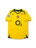Front view of Arsenal 2006 Champions League Final away shirt with yellow body, O2 sponsor, Arsenal crest, Paris final match detail, and UEFA sleeve patch.