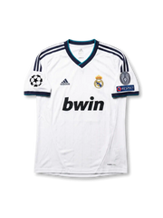 Front view of Real Madrid 2012-13 home jersey with white base, navy shoulder stripes, teal trim, club crest, sponsor, and Champions League sleeve patches.