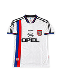 Front view of the Bayern Munich 1996-98 adidas away soccer jersey in white with vertical blue and red stripes, black polo collar, adidas Three Stripes, Opel chest sponsor, club crest, and UEFA Champions League sleeve patch.