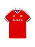 Manchester United 1984-85 home jersey, front view, red body, white V-neck collar with black trim, white adidas three-stripe shoulder detailing, adidas trefoil logo on chest, club crest, SHARP sponsor logo, striped ribbed cuffs