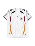 Germany 2006 home jersey, front view, white body with curved red and gold side panels, black shoulder panels, black adidas logo and stripes, three gold stars, DFB eagle crest, small tournament emblem on sleeve