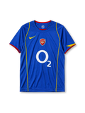 Arsenal 2004-05 away jersey, front view, blue body with yellow piping, crew-neck collar with red trim, yellow Nike swoosh, Arsenal crest, white O2 sponsor logo, yellow diagonal accent on sleeve cuff