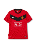 Manchester United 2010 home jersey, front view, red body with black chevron panel, black crew-neck collar, white Nike swoosh, Manchester United crest, boxed white AIG sponsor logo