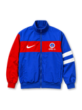 Front view of a Paris Saint-Germain 1996/97-inspired track jacket featuring a royal blue body, red horizontal chest band, red left sleeve, white stripes on the opposite sleeve, white Nike Swoosh, Opel lettering, Paris-themed crest, full zipper, and ribbed cuffs.