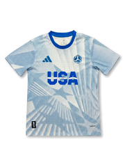 USA national team adidas 2026 FIFA World Cup pre-match jersey in white with allover blue halftone dot print, bold USA lettering at center chest, adidas logo at left, FIFA World Cup 2026 ball badge at right, royal blue crew collar, host nation badge at lower left hem
